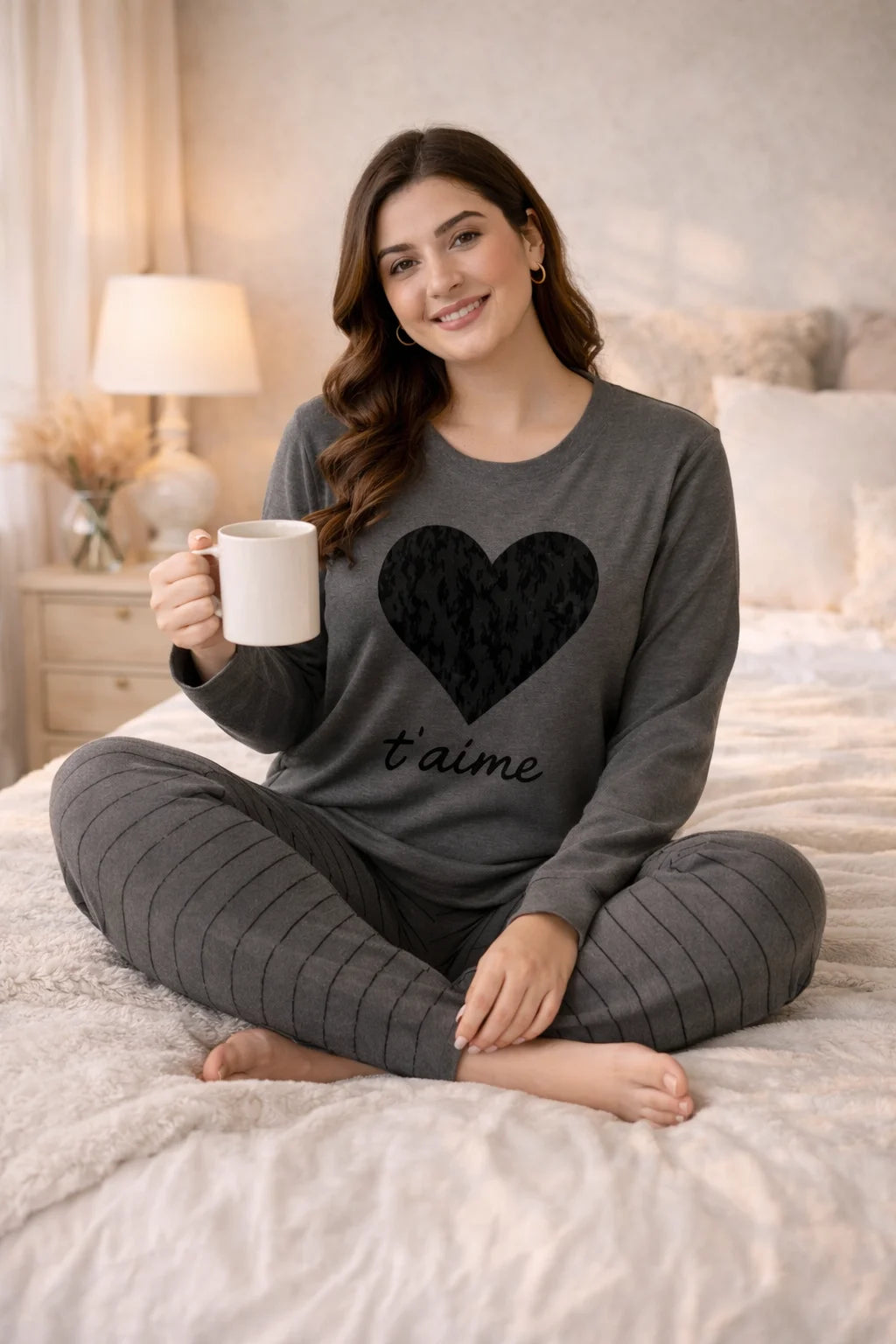 Woman in gray pajama set with heart design sitting on a bed holding a mug.
