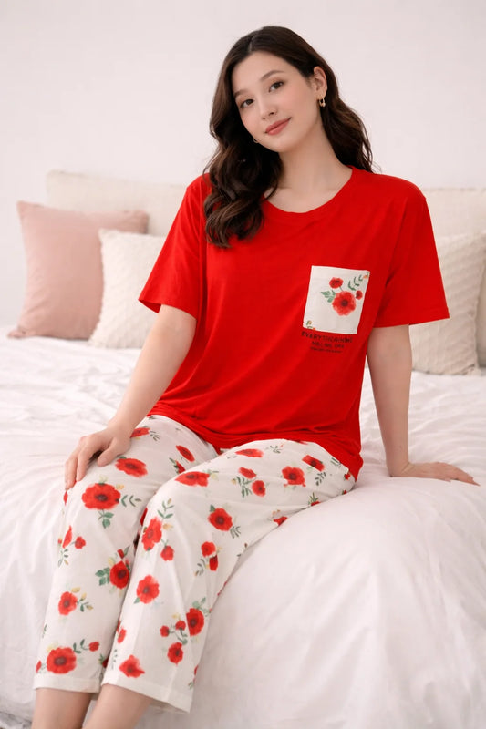 Woman wearing a red top and white pants with red floral patterns, sitting on a bed.