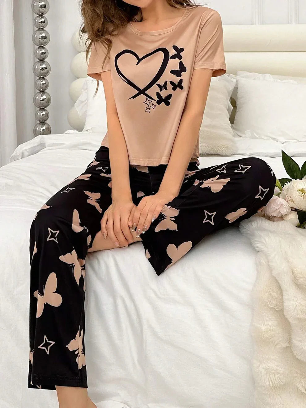 Person wearing a beige t-shirt with a heart design and black pants with butterfly patterns, sitting on a bed.