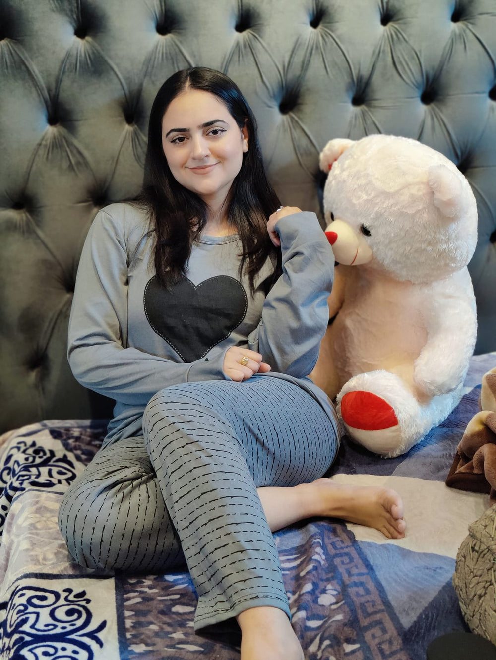 Woman sitting on a bed with a large teddy bear, wearing a heart-shaped pillow.